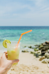 Fresh fruit cocktail with lemon slice in woman hand on the background of a sandy tropical beach. First-person view. Colorful travel and vacation concept.