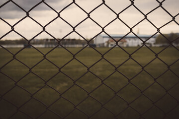 Fototapeta premium Chain link fence in farm with grassy field behind it. Cloudy sky and melancholic view.