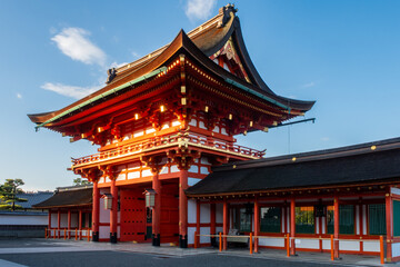 View of red entry gate to Fushimi Inari-Taisha Shinto Shrine in Kyoto, Japan, illuminated by morning sun. 