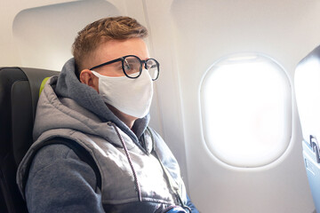 Fototapeta na wymiar Serious guy, young man on airplane, plane in glasses and medical protective sterile mask on his face traveling. Coronavirus, virus, airline concept. Pandemic covid-19. Safety in public transport