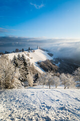 Beautiful and well known landmark Jamnik church on hilltop in wintertime. Snow covering landscape and trees. Inversion clouds in the valley. Beautiful sunny winter day. Vertical photo