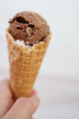 Hand holding a waffle cone with a scoop of chocolate ice cream on top of it, white background.