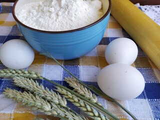 Still life of food in the kitchen eggs a bowl of white flour cookies on a plate a wooden rolling pin grains ears of wheat a bottle of sunflower oil a room flower near the window