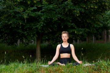 Frontal portrait of a young woman in black sportwear seated on a yoga mat in position lotus in the park, with closed eyes. Copy space.