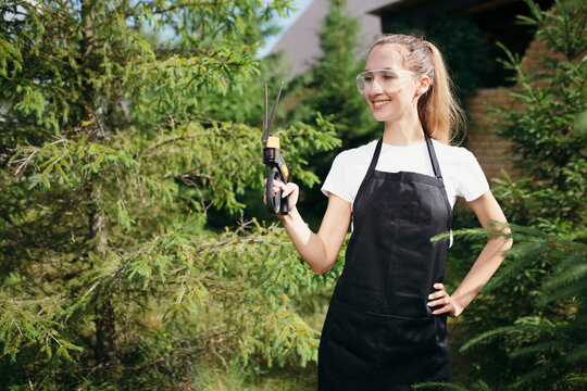 Girl gardener stands in a black apron with scissors for grass in her hands and smiles.Work in the spring on the nature.garden plot