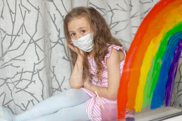 A child in a medical mask looks out the window through a painted rainbow on glass. A flash mob, a child draws a rainbow on a window pane during world quarantine due to coronavirus infection..