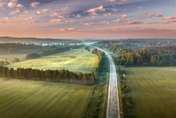 Highway going through meadow in mist surrounded by forest. Aerial view over picturesque landscape in fog at sunrise. 
 - obrazy, fototapety, plakaty