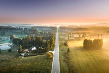 Highway going through meadow in mist surrounded by forest. Aerial view over picturesque landscape in fog at sunrise. 
