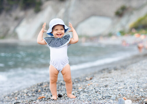 Cute Baby Girl Standing On The Sea Coast In A White Swimsuit And Laughting