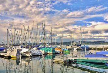 Fototapeta na wymiar Sailboat harbor on Lake Constance, in the background you can see the Lake Constance ferry. The sky has many white clouds on blue, in the water you can see the mirrored boats.