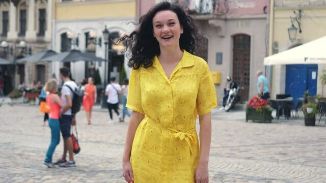 Attractive girl wearing yellow dress filming selfie video. Curly woman having video stream on holiday, waving at webcam on smartphone camera sharing travel vacation. Urban city background.