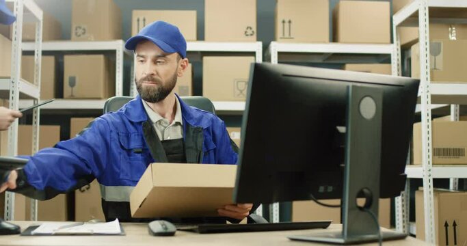 Handsome Caucasian mailman in blue iniform and cap sitting at table in postal office store and working at computer. Male post courier filling in invoice and entering data while scanning bar code.