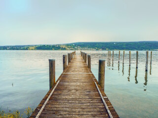 A jetty of wooden planks protrudes into Lake Constance (Germany). The almost light turquoise evening sky is reflected in the water, on the opposite side you can see a strip of land.