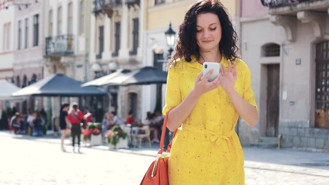Young woman wearing yellow dress with red handbag walking around old street using smartphone chatting. Communication, social networks, online shopping concept.