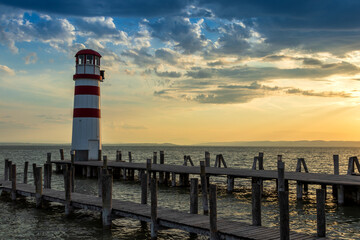 A wooden pier leading to the lake and a red and white lighthouse on Lake Neusiedl in Podersdorf, Austria. In the background is a dramatic sky at sunrise.