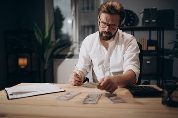 Bearded businessman in eyeglasses and white shirt counting money cash that holding in hands while sitting at desk. Mature man dividing dollars into different piles at modern office.