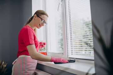 Attractive woman with blond hair wearing eyeglasses, apron and rubber gloves, cleaning windowsill with cloth and detergent. Concept of household duties.