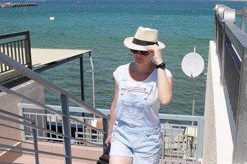 Tourist girl in denim overalls, hat and sunglasses holds her hat and climbs the stairs from the sea promenade