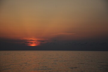 The rays of the setting sun behind small dark clouds over a calm sea