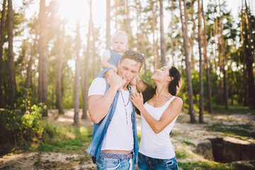 Family mom, dad and daughter sits at the daddy on the shoulders, and parents kiss on the nature in the forest in the summer at sunset