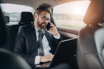 Concentrated businessman in eyewear and black suit having conversation on mobile phone while riding in car. Mature man sitting on backseat with opened laptop on knees.