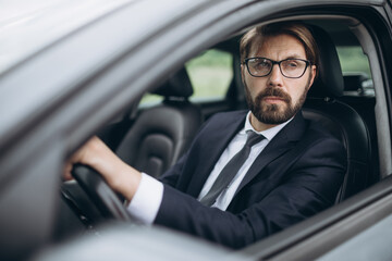 Portrait of confident bearded man wearing eyeglasses and black suit driving own luxury car. Successful businessman is hurry to important meeting.