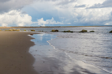 Ocean Wave on Sandy Beach with Large Rocks and Sun Peeking Through Clouds