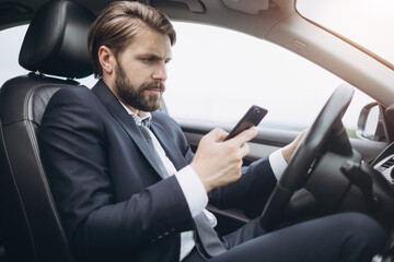 Fototapeta na wymiar Side view of busy bearded man dressed in formal clothing using smartphone while driving car. Mature businessman texting message while sitting in automobile.