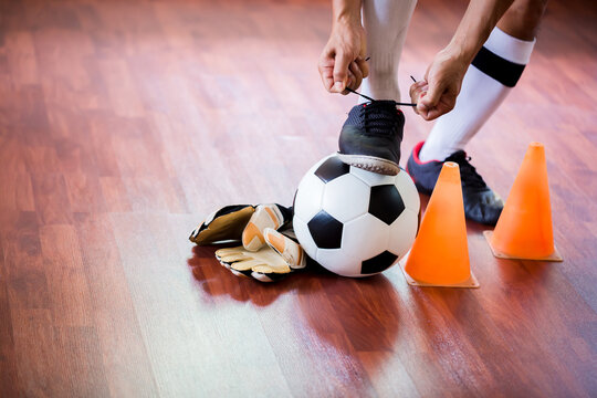 Soccer ball with futsal player sitting and tying sport shoelaces in stadium hall. Futsal player and training equipment on the wooden floor.