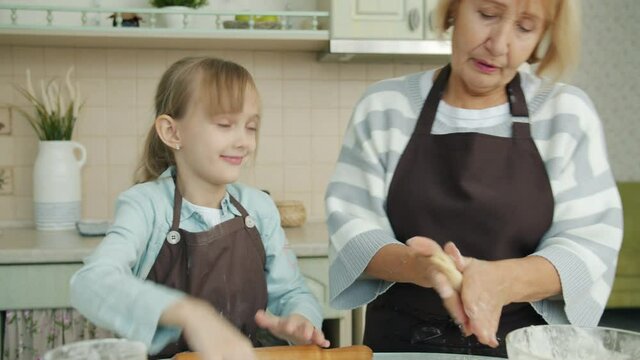 Joyful kid wearing apron is helping grandma in kitchen rolling dough then having fun touching granny's nose with finger covered with flour and laughing