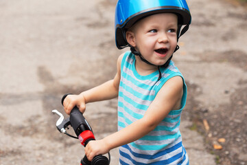 Happily smilling toddler boy in blue shirt with stripes and helmet standing on the road with...
