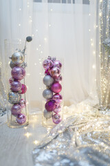Purple bulbs in transparent glass vase. Christmas and New Year decorations and toys. Blurry white background with warm garland lights and bokeh. Winter magic. Place for text. Vertical shot.