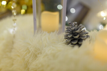 Silver painted pine cone on Merry Christmas and Happy New Year background. Candle standing on white fur carpet. Blurry warm garland lights on background. Winter holidays greeting card, copy space.