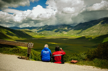 Fototapeta na wymiar Biker couple admiring spectacular mountain scenery: panorama of the Piano Grande during the flowering of the fields, near Castelluccio di Norcia (Umbria, Italy), in the Sibillini mountains.
