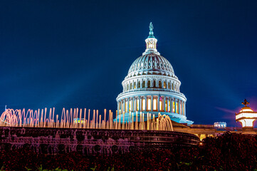Marble dome of the United States Capitol building illuminated by rays of architectural light on a clear blue night with fountain with lights in foreground in Washington DC