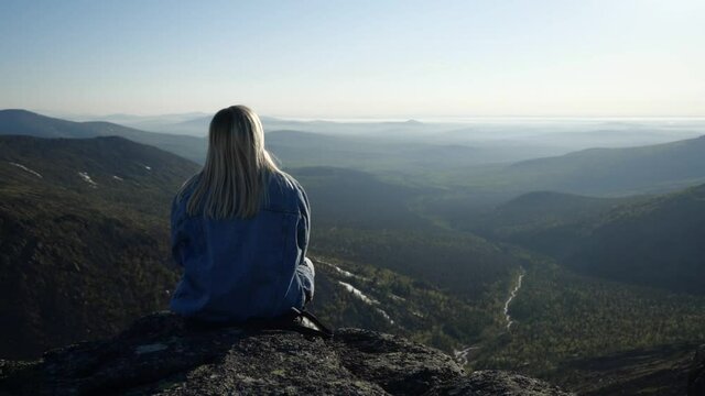 A woman with blond hair in a blue denim jacket sits on the edge of a cliff with her back to the camera, and looks out over the mountain valley under the sun