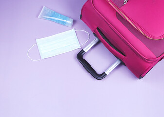 COVID-19 protection while traveling and new normal concept.Top view or flat lay of pink suitcase  with surgical face mask and alocohol sanitizer gel  on purple background