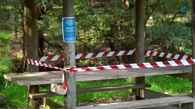 Wooden table at a picnic area is closed due to covid-19 restrictions. A sign on a pole states about social distancing. Red and white tapes block the area.