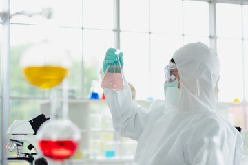 Asian scientist man in personal protective equipment uniform wearing glasses protection, mask and green glove sitting and holding liquid sample in glass conical to look analysis in laboratory