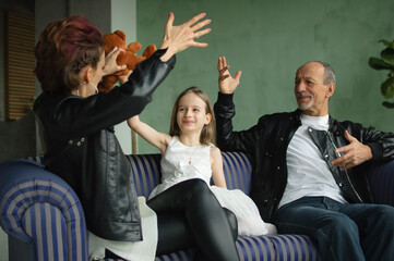 Family portrait of adult daughter, little granddaughter and senior grandfather in loft room with houseplants. Man and woman are wearing black leather jackets in punk style, generation concept