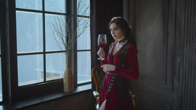 Beautiful young woman queen holding glass bloody wine in hands. Lady Brunette long wavy hair. Red sexy carnival vampire dress costume. dark black interior gothic medieval room, window. Fashion model