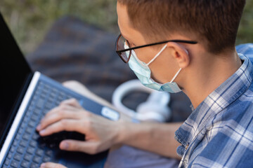 guy in an antiviral mask in a park on a plaid is working on a laptop