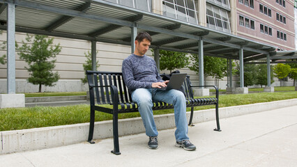 High quality picture of a young men who is sitting on a train station and working on his laptop which is holding on the lap. Young men sitting on a black metal banch on a station in the city. 