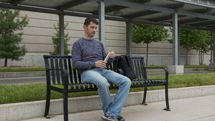 High quality picture of a young man who is sitting on a metal bench outside of train and bus station and drinking water from the stainless steel thermos which he took from the backpack.