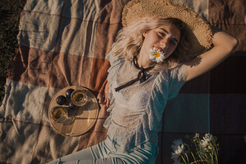 young curly blonde in a straw hat with glasses with daisies in a basket lies on a plaid top view