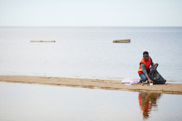 African young man picking up the garbage into bags he cleaning the bank of the river outdoors