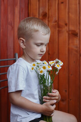 little boy in a white T-shirt sits on a chair against a wooden wall and holds in his hands a glass vase with a bouquet of chamomiles, looks down.
lmage with selective focus