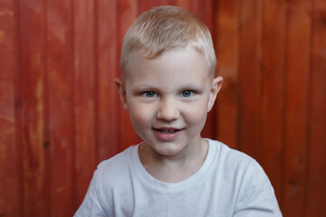 little blond boy in a white T-shirt stands on the background of a wooden wall and smiles, looks at the camera.
lmage with selective focus