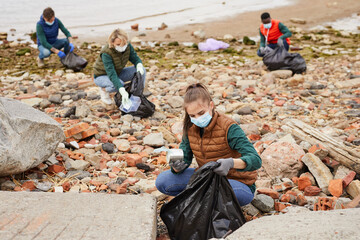 Group of young volunteers picking up the garbage in bags near the coastline outdoors