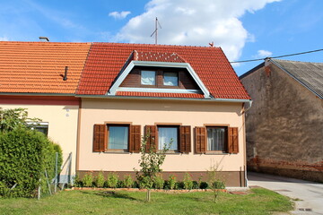 Attached completely renovated small suburban family house with new wooden frame windows and open window blinds surrounded with old family houses and flowers in front on cloudy blue sky background